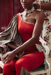 A person sits wearing an elegant red, one-shoulder gown embellished with sparkly details. The fabric appears luxurious with an additional striped material draped over the shoulder. A large, intricate floral choker adorns the neck, and another person is seen helping adjust the gown.