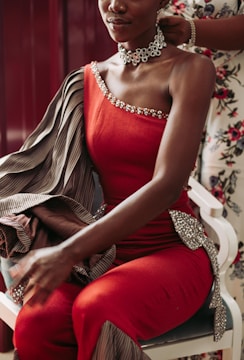 A person sits wearing an elegant red, one-shoulder gown embellished with sparkly details. The fabric appears luxurious with an additional striped material draped over the shoulder. A large, intricate floral choker adorns the neck, and another person is seen helping adjust the gown.