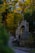 A tranquil cemetery scene with tombstones and ornate mausoleums lining a curving path. The area is surrounded by lush green foliage with sunlight filtering through the trees, casting a serene glow.