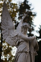 A detailed stone statue of an angel with large, intricately carved wings is depicted. The angel appears serene and contemplative, gazing downward while holding an orb close to its chest. The background features blurred trees, suggesting an outdoor setting, possibly a park or cemetery.