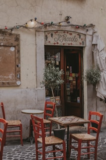 A quaint outdoor seating area of a small cafe with red wooden chairs and square tables on a cobblestone street. The cafe sign reads 'Il Piccolo' and above the entrance are string lights with colorful bulbs. Potted plants flank the entrance, creating a cozy and inviting atmosphere.
