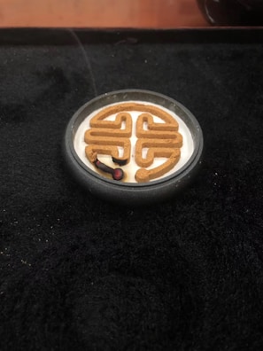 An incense coil is burning in a decorative pattern placed in a small, round container. The surrounding surface is dark, creating a contrast with the light-colored incense ash and the smoke rising gently.