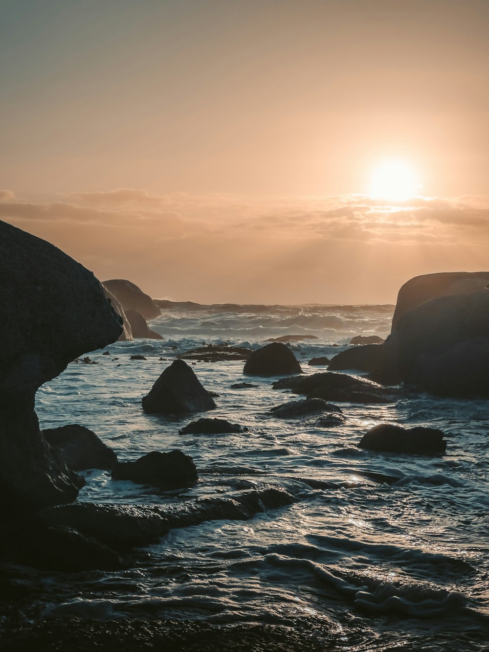 the sun is setting over the ocean with rocks in the foreground