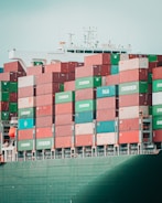 A large cargo ship is stacked high with multicolored shipping containers labeled 'Evergreen,' 'Italia,' and others. The containers are arranged in neat rows on the deck of the vessel. The structure of the ship's command tower is visible behind the containers, with various antennas and a small flag.