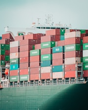 A large cargo ship is stacked high with multicolored shipping containers labeled 'Evergreen,' 'Italia,' and others. The containers are arranged in neat rows on the deck of the vessel. The structure of the ship's command tower is visible behind the containers, with various antennas and a small flag.