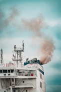 A large ship emits thick smoke from its smokestack. The ship features complex structures and antennas on its upper deck, with visible company branding. The sky is overcast, providing a muted backdrop.