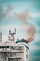 A large ship emits thick smoke from its smokestack. The ship features complex structures and antennas on its upper deck, with visible company branding. The sky is overcast, providing a muted backdrop.