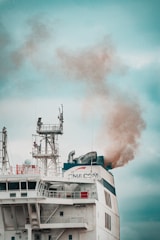 A large ship emits thick smoke from its smokestack. The ship features complex structures and antennas on its upper deck, with visible company branding. The sky is overcast, providing a muted backdrop.