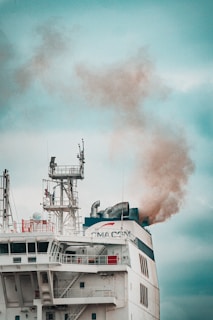 A large ship emits thick smoke from its smokestack. The ship features complex structures and antennas on its upper deck, with visible company branding. The sky is overcast, providing a muted backdrop.