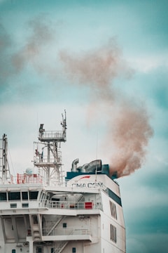 A large ship emits thick smoke from its smokestack. The ship features complex structures and antennas on its upper deck, with visible company branding. The sky is overcast, providing a muted backdrop.
