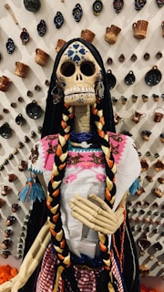 A decorative skeleton figure is adorned with vibrant clothing and long braided hair against a backdrop of various ceramic mugs and wall decorations. The skull features intricate blue patterns and the figure wears a colorful traditional outfit with bright pink, orange, and blue tones.