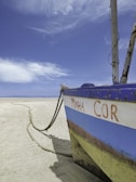 A weathered, beached boat with the words 'MINHA COR' is positioned on a sandy beach. It is tied with a thick rope that extends along the sand. The sky is vast and blue with wispy clouds, creating a serene, open atmosphere.