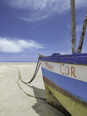 A weathered, beached boat with the words 'MINHA COR' is positioned on a sandy beach. It is tied with a thick rope that extends along the sand. The sky is vast and blue with wispy clouds, creating a serene, open atmosphere.
