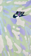 A colorful fabric pattern featuring abstract shapes in soft pastel hues of green, white, and purple. The image includes a logo with text and a small graphic element overlaid on the pattern.