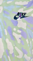A colorful fabric pattern featuring abstract shapes in soft pastel hues of green, white, and purple. The image includes a logo with text and a small graphic element overlaid on the pattern.