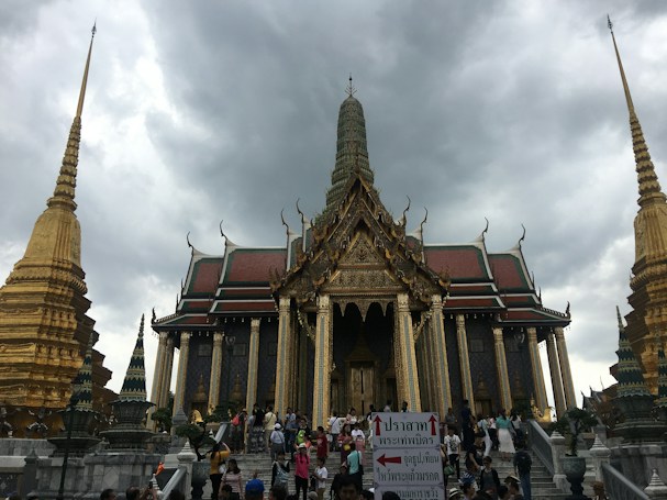 A large, ornate temple with intricate architecture featuring spires and a richly decorated roof stands against a backdrop of cloudy skies. The elaborate design includes gold detailing and vibrant colors. People are gathered at the entrance, walking up the stairs in front of the temple. Decorative structures and sculptures are visible surrounding the temple.