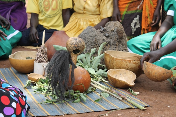 A group of people are seated on the ground, surrounding a ceremonial arrangement that includes traditional gourds, leaves, seeds, and a woven mat. The setting appears to be outdoors, with the participants dressed in colorful garments.