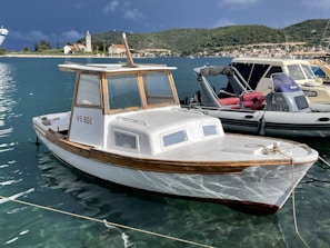A small wooden boat with the registration number VS 308 is docked on a calm body of water. The boat has a cabin with large windows and a white hull with wooden trim. In the background, there is a scenic view of a town with a prominent church and lush green hills. Several other boats are also docked nearby, including a gray inflatable boat with a red interior.