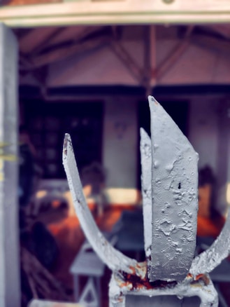 A worn and rusted metal gate is prominently in focus, displaying textured and chipped gray paint. The background is blurred, showing the interior of an outdoor area with an open roof structure, possibly part of a patio or porch.
