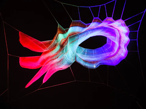 A luminous sculpture with a swirling abstract design, illuminated in vibrant shades of red, blue, and pink, set against a dark background. The installation appears to be made of net-like material, creating a mesmerizing, holographic effect.