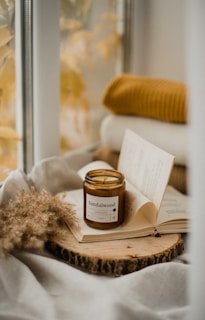 A cozy setting with a lit sandalwood-scented candle placed on an open book, resting on a wooden slab. The background includes a softly blurred scene with autumn leaves and a folded mustard-colored blanket, conveying a warm and inviting atmosphere.