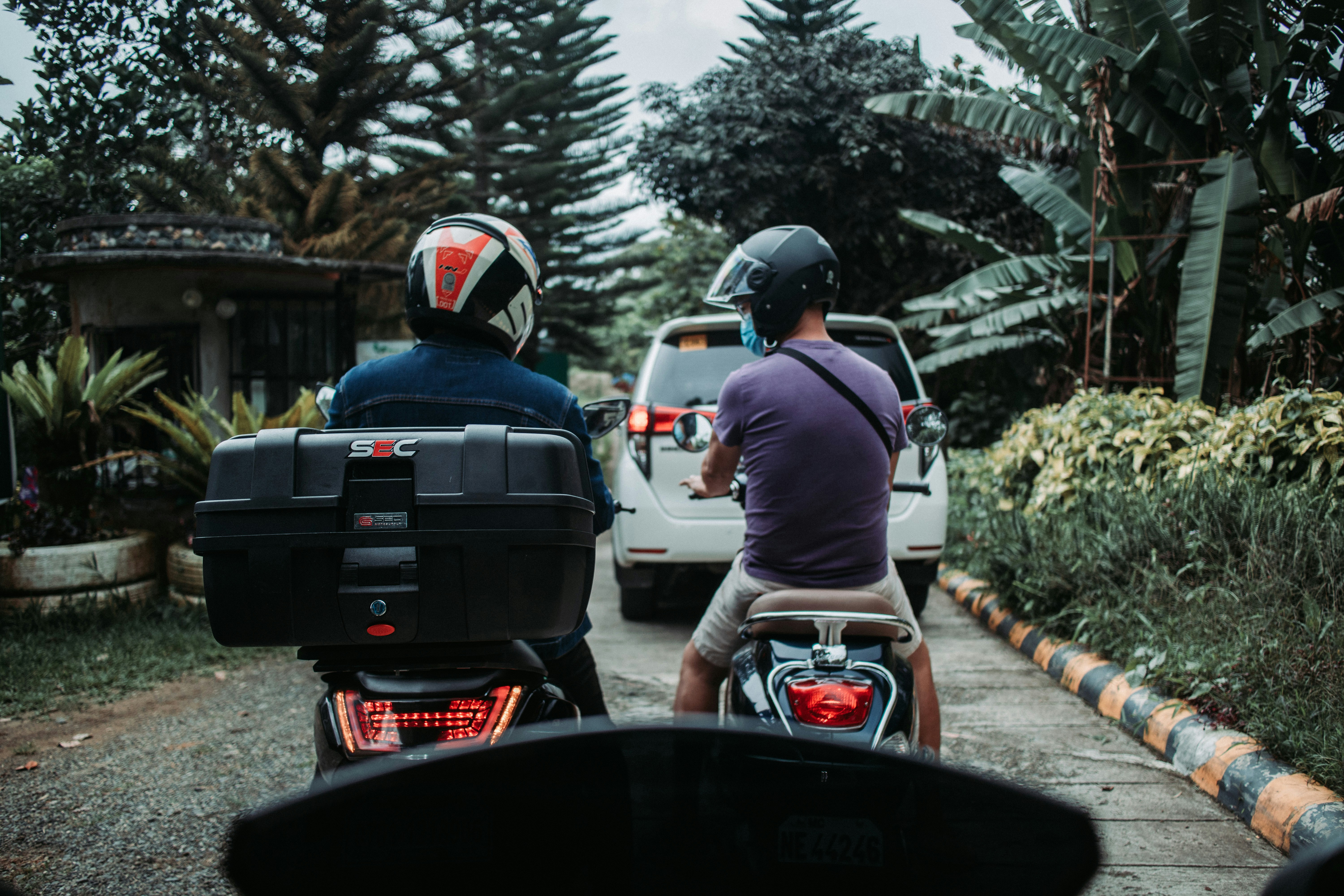 Dua pengendara skuter tampak dari belakang, salah satunya dengan top box hitam, di jalan berpepohonan