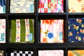 Various squares of colorful fabric are neatly displayed in a black grid. Each fabric features a unique pattern, including floral designs, geometric shapes, and prints with small owl illustrations. The colors range from vibrant reds and blues to muted greens and yellows.