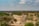 A vast African landscape displays a dry riverbed surrounded by dense green vegetation and scattered trees. A solitary vehicle traverses the dusty path, suggesting a safari experience. The sky is clear with a few scattered clouds, presenting a serene and open ambiance.