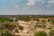 A vast African landscape displays a dry riverbed surrounded by dense green vegetation and scattered trees. A solitary vehicle traverses the dusty path, suggesting a safari experience. The sky is clear with a few scattered clouds, presenting a serene and open ambiance.