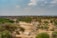 A vast African landscape displays a dry riverbed surrounded by dense green vegetation and scattered trees. A solitary vehicle traverses the dusty path, suggesting a safari experience. The sky is clear with a few scattered clouds, presenting a serene and open ambiance.