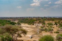 A vast African landscape displays a dry riverbed surrounded by dense green vegetation and scattered trees. A solitary vehicle traverses the dusty path, suggesting a safari experience. The sky is clear with a few scattered clouds, presenting a serene and open ambiance.