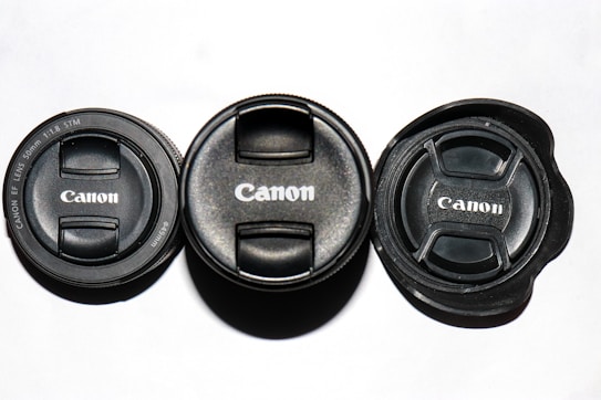 Three Canon lens caps are arranged in a row on a white surface, showcasing the brand's logo prominently. The caps vary slightly in design, indicating different lens models.