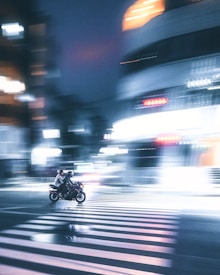 A motorcyclist with a passenger is riding through a blurred urban street at night. The city lights create a dynamic and fast-moving effect, with vibrant light streaks and reflections on the wet road surface.