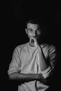 A monochromatic portrait of a person wearing a long-sleeved shirt with one arm crossed and the other hand resting on their chin. The lighting is dramatic with shadows emphasizing the facial features and clothing texture against a black background.
