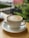 A frothy coffee is served in a white ceramic cup on a matching saucer, resting on a wooden table. The background is soft-focused, suggesting green foliage and blurred outdoor scenery, contributing to a tranquil atmosphere.