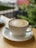 A frothy coffee is served in a white ceramic cup on a matching saucer, resting on a wooden table. The background is soft-focused, suggesting green foliage and blurred outdoor scenery, contributing to a tranquil atmosphere.