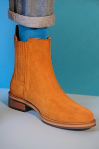 A brown suede ankle boot with a classic design is worn with rolled-up jeans and bright blue socks. The background is a solid teal color, creating a stylish and fashionable composition.