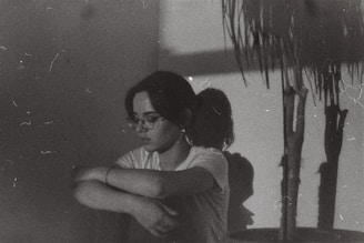 A person with glasses and a white shirt sits against a wall, arms wrapped around their knees. The room is dimly lit, casting shadows on the wall. A plant is visible to the right, with its shadow also cast on the wall. The overall aesthetic suggests a melancholic or introspective mood.