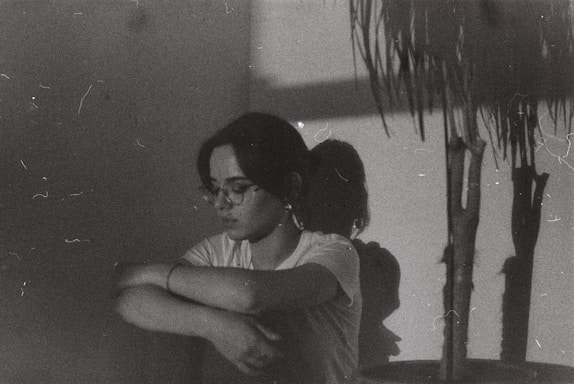 A person with glasses and a white shirt sits against a wall, arms wrapped around their knees. The room is dimly lit, casting shadows on the wall. A plant is visible to the right, with its shadow also cast on the wall. The overall aesthetic suggests a melancholic or introspective mood.