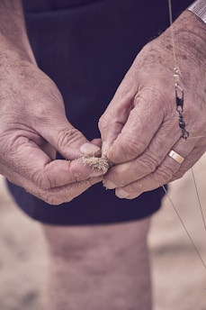 Close-up of a pair of elderly hands holding and examining fishing bait with sand, possibly preparing it for use. The hands appear weathered, suggesting experience in fishing or outdoor activities. A fishing line and hook are partially visible.