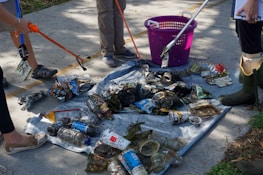 A group of people are using grabbers to collect litter, including plastic bottles, cans, and miscellaneous trash, on a tarp placed on a concrete surface. A pink basket is nearby, and some individuals are wearing rubber boots and casual shoes.