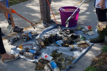 A group of people are using grabbers to collect litter, including plastic bottles, cans, and miscellaneous trash, on a tarp placed on a concrete surface. A pink basket is nearby, and some individuals are wearing rubber boots and casual shoes.