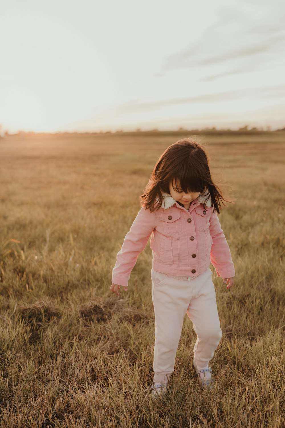 a little girl in a pink jacket and white pants in a field