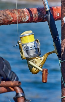 A close-up view of a fishing reel attached to a fishing rod. The reel has a gold body with a wooden handle and yellow fishing line wound around it. The fishing rod is attached to a rusted, red-brown metal railing. The background shows out-of-focus blue water, indicating that the scene is likely by a body of water.