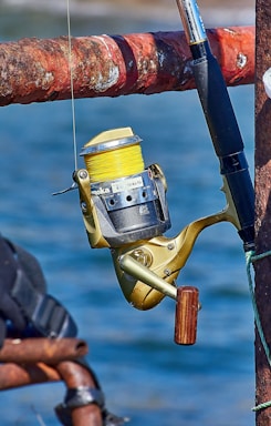 A close-up view of a fishing reel attached to a fishing rod. The reel has a gold body with a wooden handle and yellow fishing line wound around it. The fishing rod is attached to a rusted, red-brown metal railing. The background shows out-of-focus blue water, indicating that the scene is likely by a body of water.