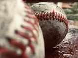 Two weathered baseballs with visible red stitching sit on a wet surface, with one in focus and the other blurred in the foreground. The texture of the balls is rough, suggesting frequent use.