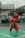 A person dressed in a traditional red martial arts uniform is performing a kata or martial arts routine on a green sports court. The uniform includes a patterned sarong around the waist and a black head covering. There are spectators in the background, mostly school-aged children, watching behind a fence. The scene is outdoors, and the sky appears overcast.