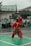 A person dressed in a traditional red martial arts uniform is performing a kata or martial arts routine on a green sports court. The uniform includes a patterned sarong around the waist and a black head covering. There are spectators in the background, mostly school-aged children, watching behind a fence. The scene is outdoors, and the sky appears overcast.
