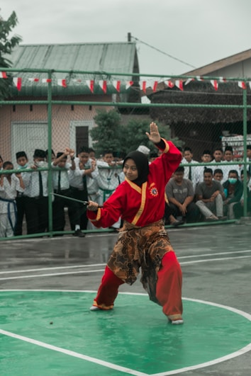 A person dressed in a traditional red martial arts uniform is performing a kata or martial arts routine on a green sports court. The uniform includes a patterned sarong around the waist and a black head covering. There are spectators in the background, mostly school-aged children, watching behind a fence. The scene is outdoors, and the sky appears overcast.
