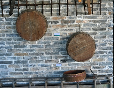 An arrangement of rustic wooden tools and objects mounted against a gray brick wall. Two large wooden circular sieves or lids hang on the wall, with a third similar item placed horizontally on a shelf or rack near the bottom. Nearby, there are additional wooden tool handles and metal implements displayed.